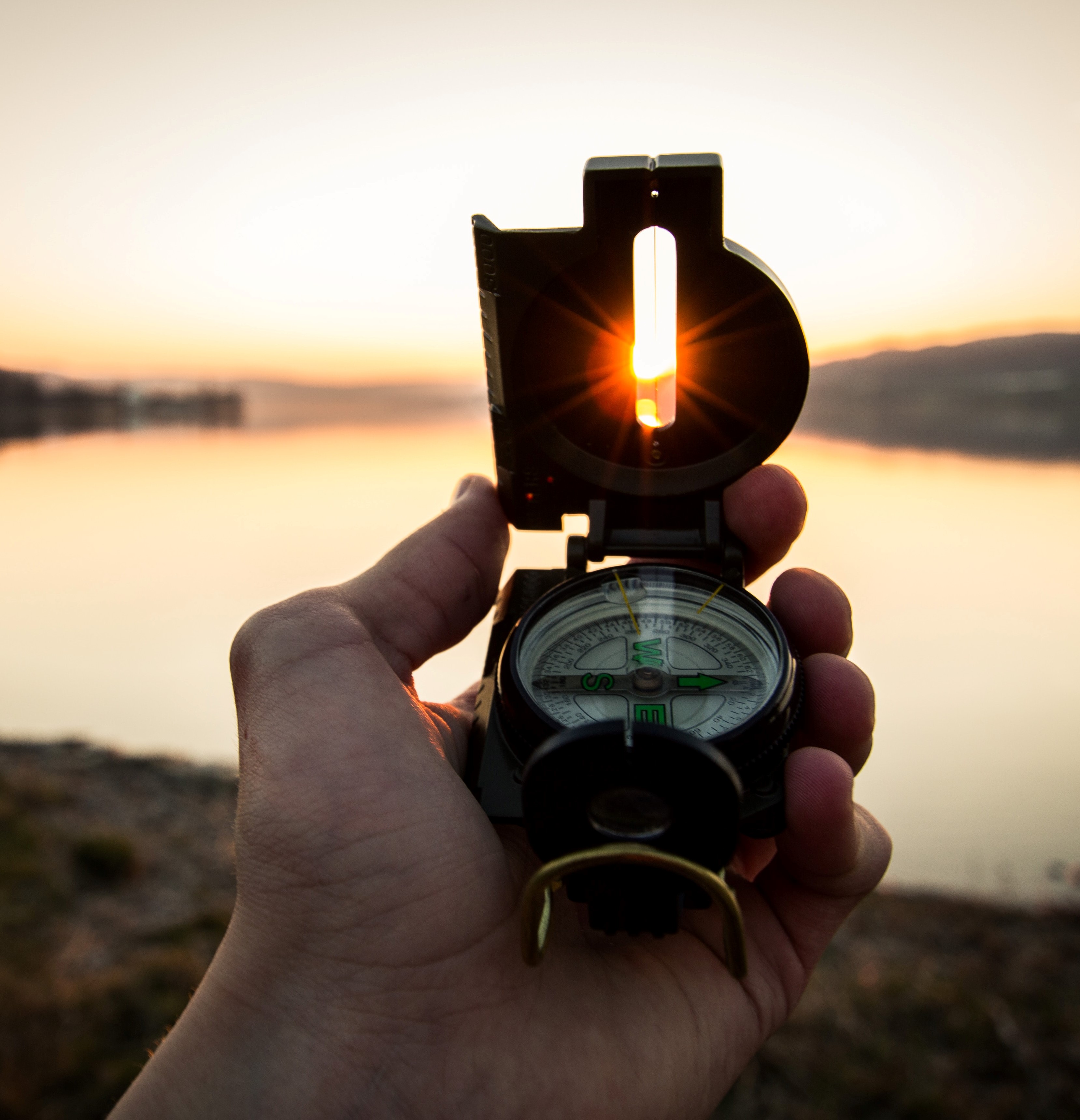 Person holding a compass at sunrise on a still lake, symbolizing the importance of seeking direction and purpose in being a Protopian.